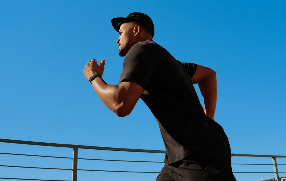 An athlete wearing a black t-shirt and cap sprinting outdoors under a blue sky while testing a screenless fitness tracker on his wrist.