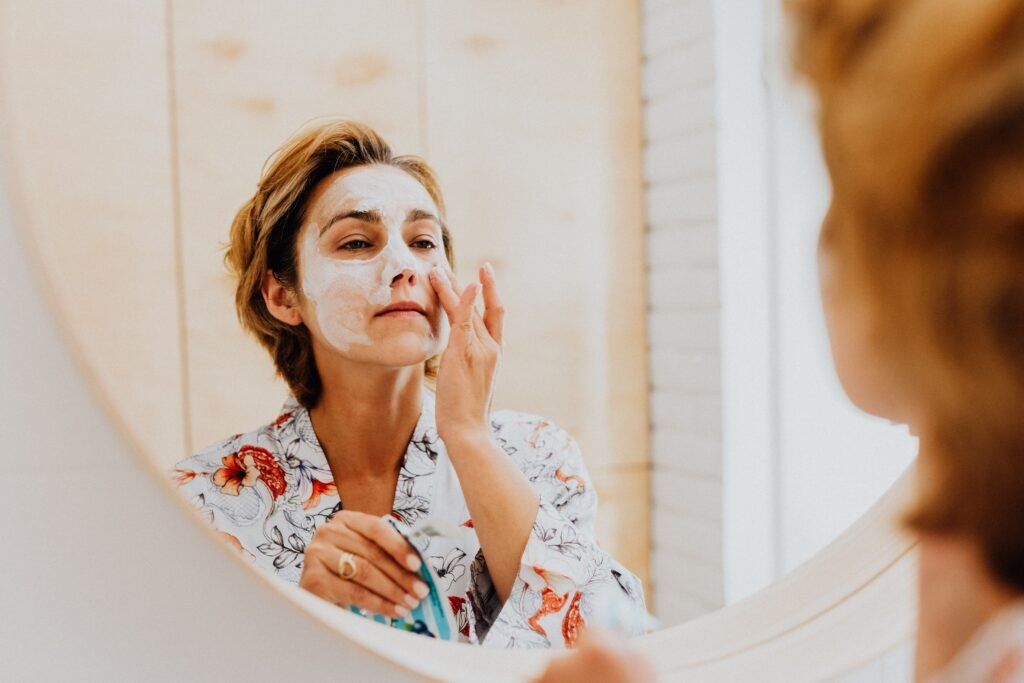 A woman looking in a bathroom mirror while applying a white cream face mask as part of a skincare routine by skin type.